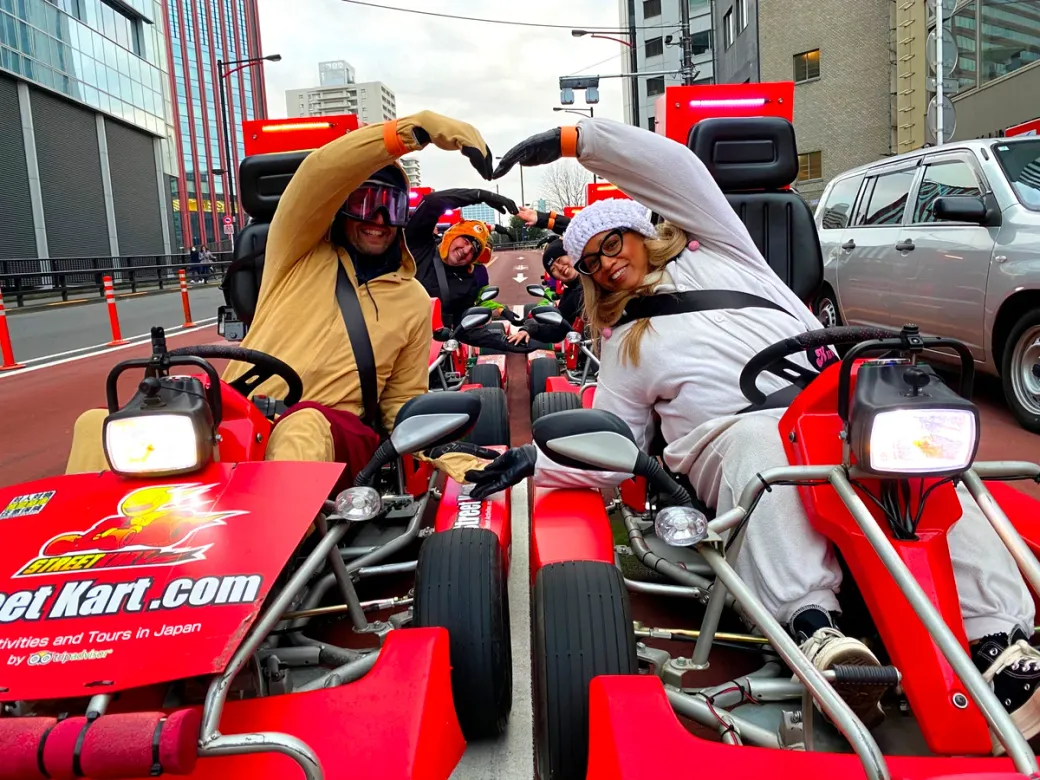 Two people in red go-karts on a city street form a heart shape with their arms, smiling at the camera in colorful costumes and helmets.