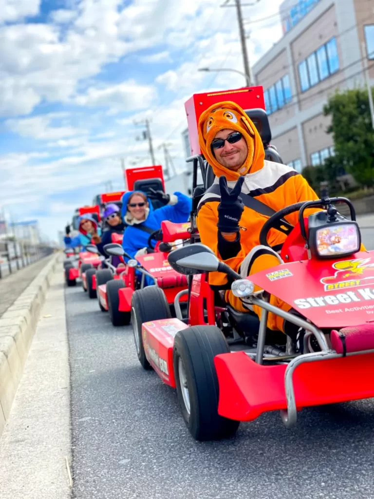 IMG_2813 - StreetKart Person in an orange mascot suit on a red go-kart, giving a peace sign in a lineup of riders on a city street.