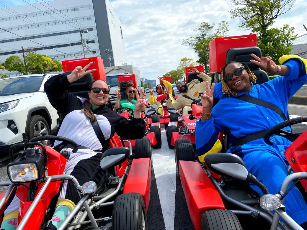 Group of people riding red go-karts on a city street, smiling and flashing peace signs at the camera during an outdoor ride.