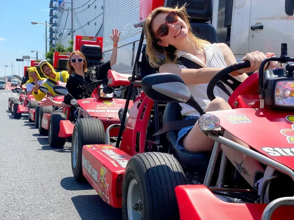 Group of people in red go-karts lined up on a sunny city street, smiling and waving at the camera.
